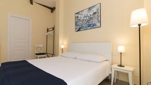 A hotel room with a bed, a nightstand with a lamp, a chair, a coat rack, and a painting on the wall. - Court of Nobles (Conversano)