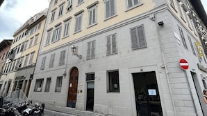 Un immeuble à plusieurs étages, de style architectural classique, avec une entrée centrale dotée d’une porte en bois, de nombreuses fenêtres avec volets et une rue pavée en pierre devant.