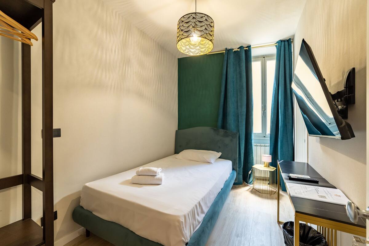 A neatly made bed with a white comforter and a folded towel, a desk with a lamp and papers, and a window with curtains.