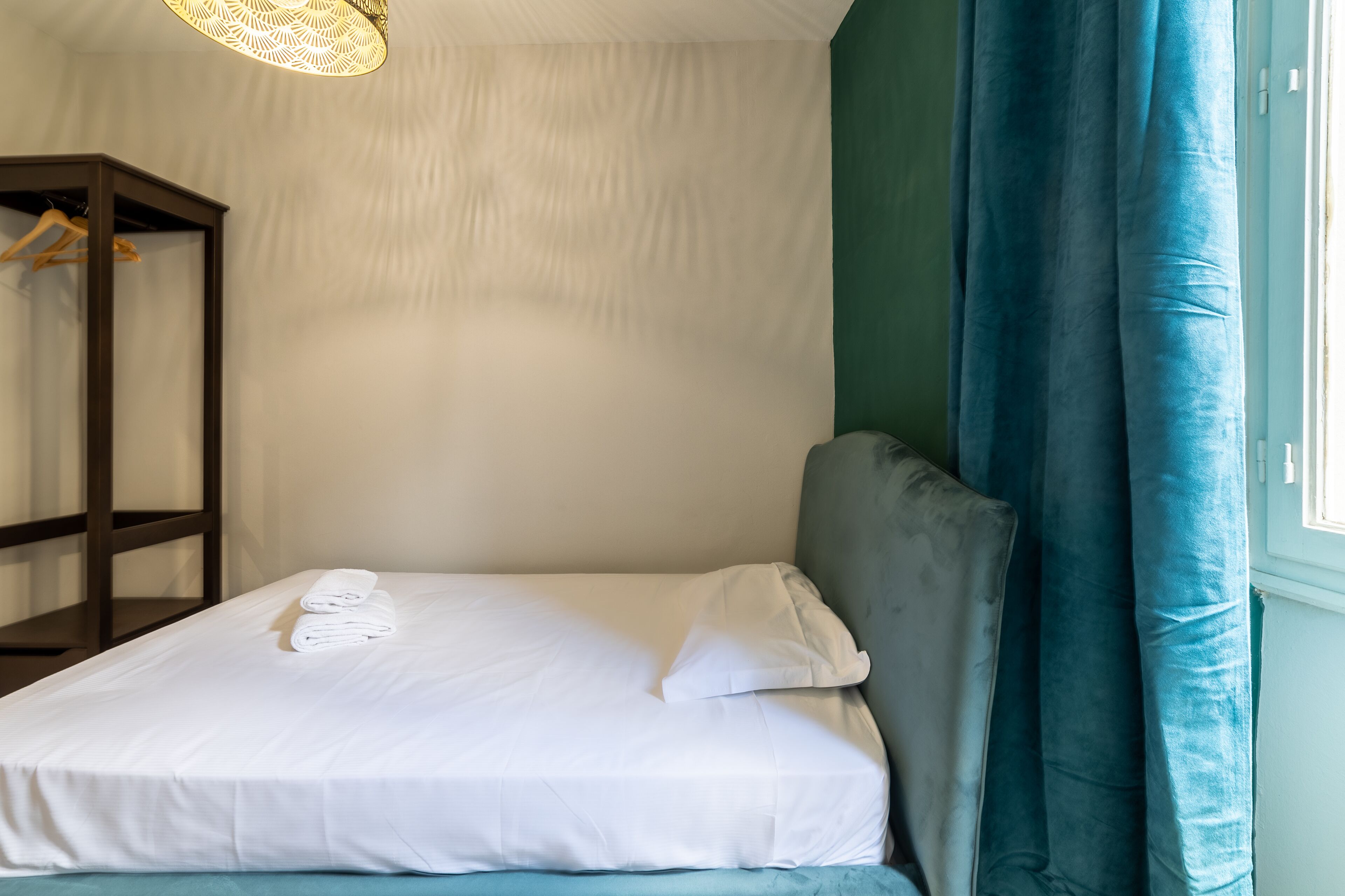 A neatly made bed with white linens and pillows, a wooden wardrobe, and a window with blue curtains.