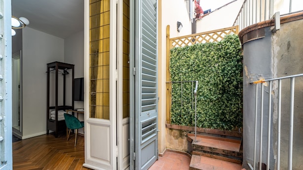 Un couloir avec un plancher en bois, une porte et un escalier menant à un balcon bordé d’une haie verte.