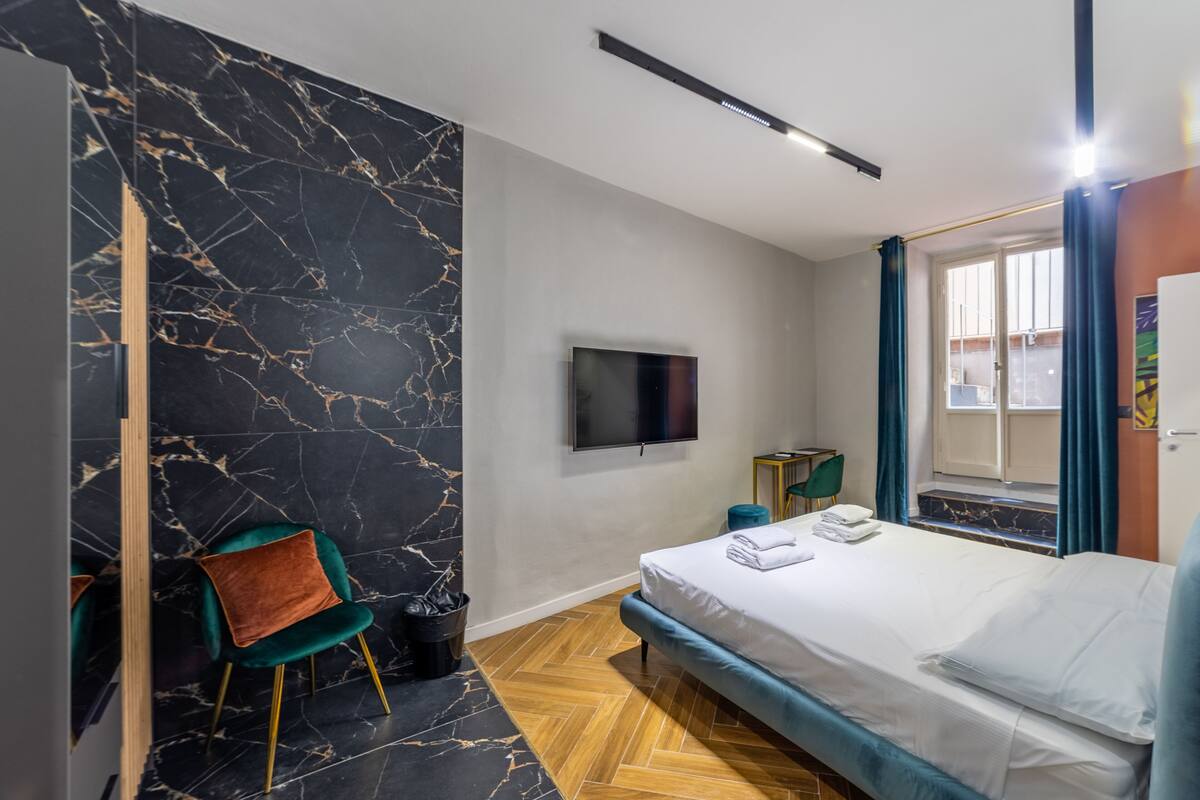 A modern hotel room with a marble wall, a bed with a blue headboard, a flat-screen TV, a wooden floor, and a window with curtains.