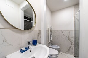 A modern bathroom with a white sink, a round mirror, a bidet, and a glass-enclosed shower.