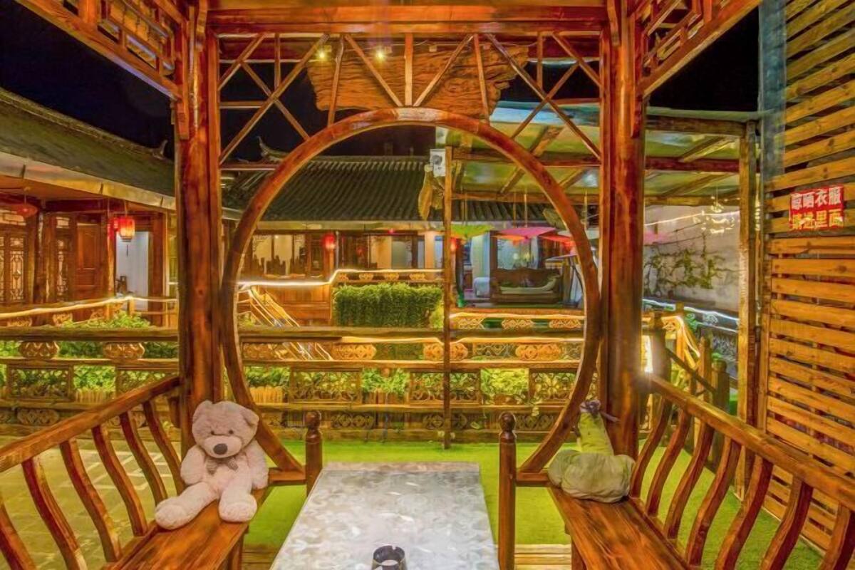 A wooden gazebo with a swing, a teddy bear on one of the benches, and a view of a traditional building in the background.
