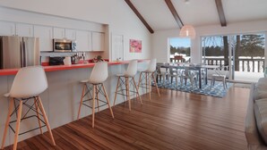 Een moderne keuken met witte kastjes, een roestvrijstalen koelkast en een rode aanrecht. Drie witte barstoelen en een eettafel met stoelen staan er. De kamer heeft houten vloeren en een groot raam met uitzicht op bomen.