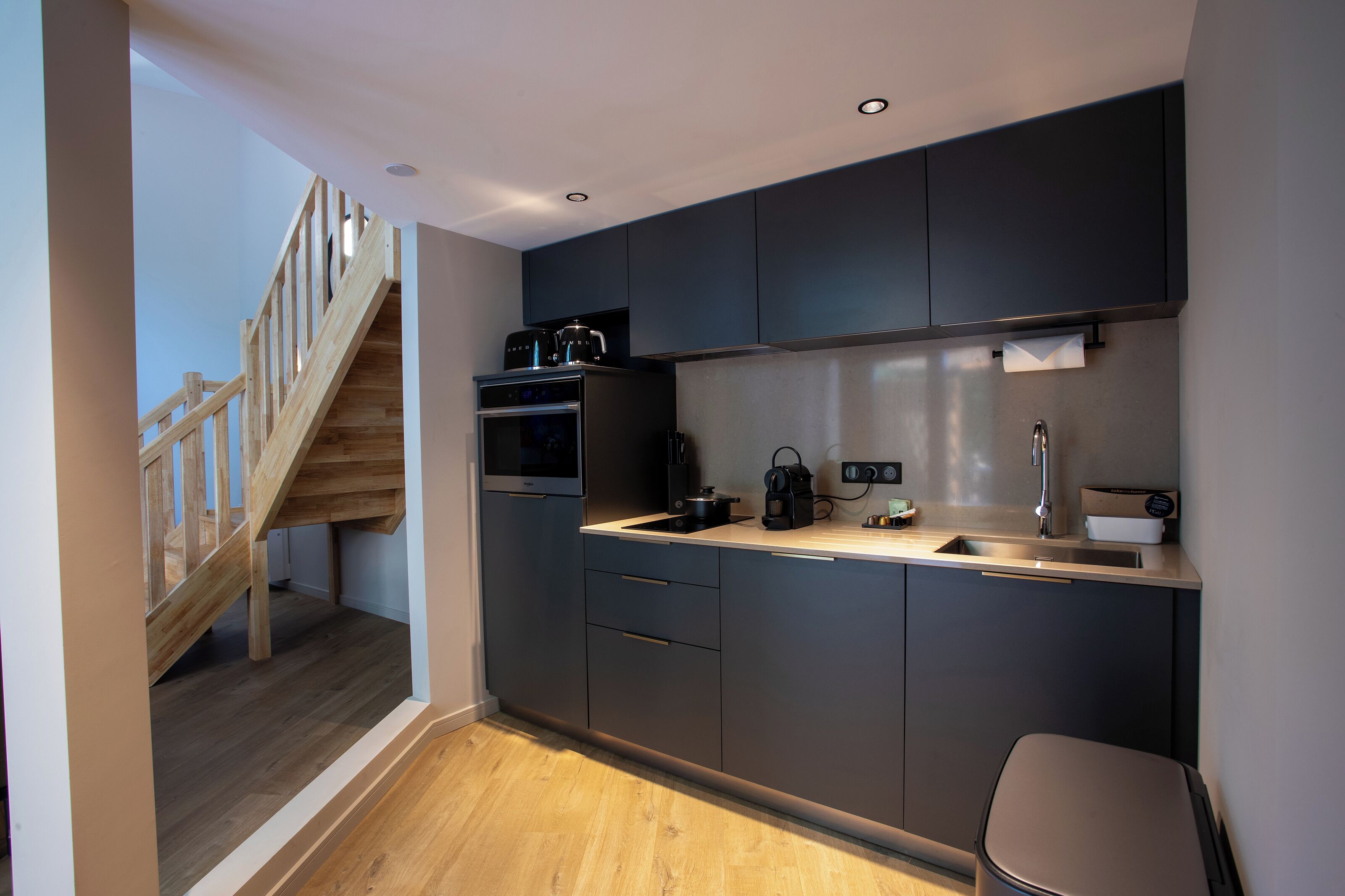 Una cocina moderna con armarios oscuros, una escalera de madera y un lavamanos de acero inoxidable.