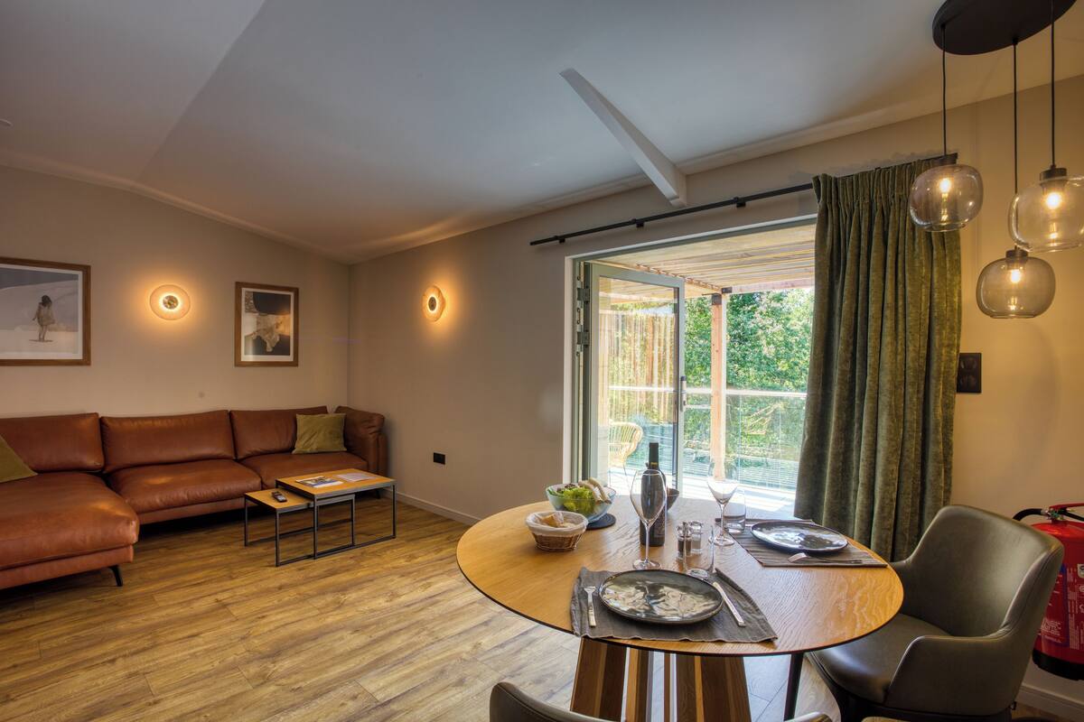 A modern living room with a sofa, a dining table set for two, and a large window with curtains.