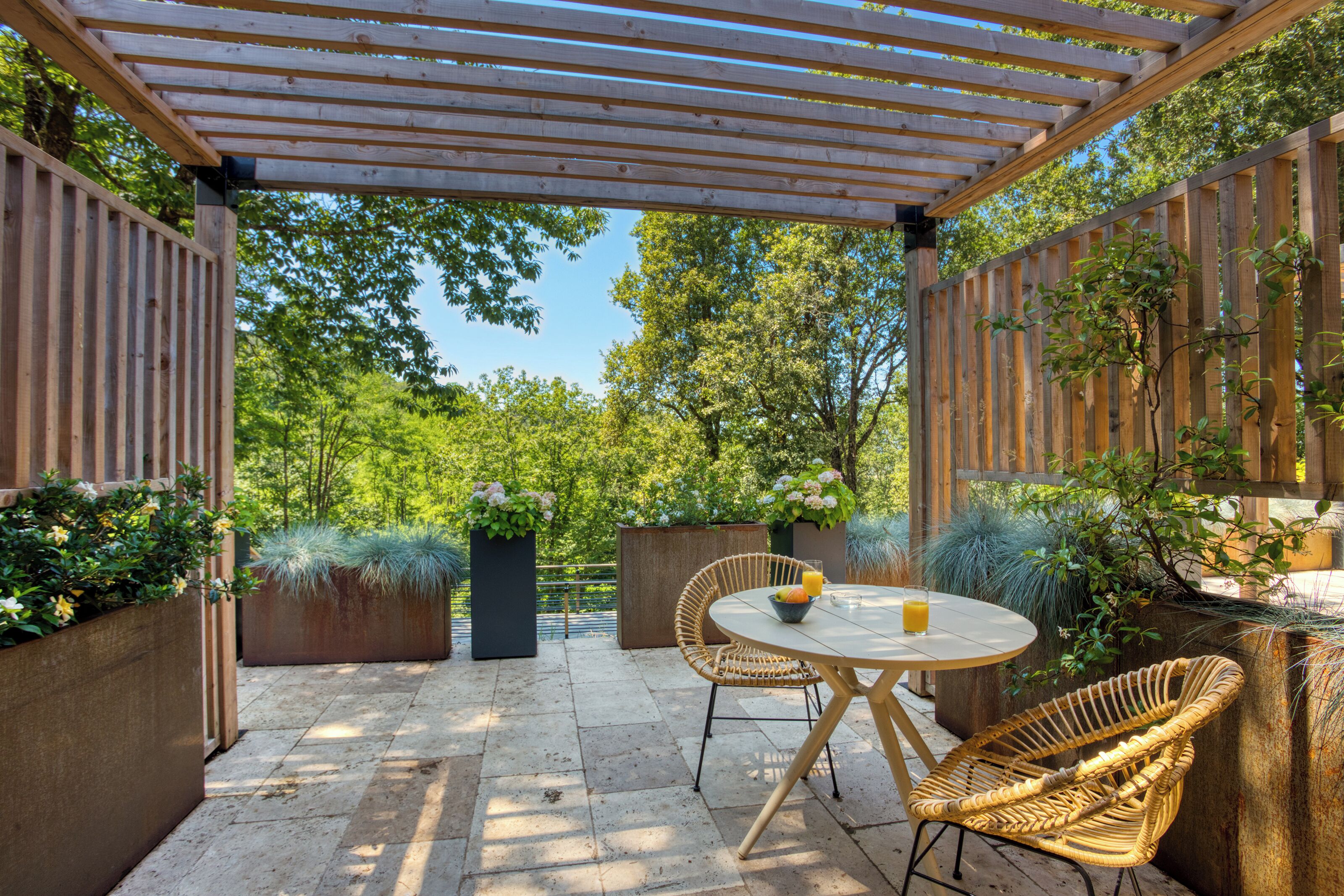 Eine Terrasse mit einer Pergola, Korbstühlen, einem runden Tisch und Topfpflanzen.
