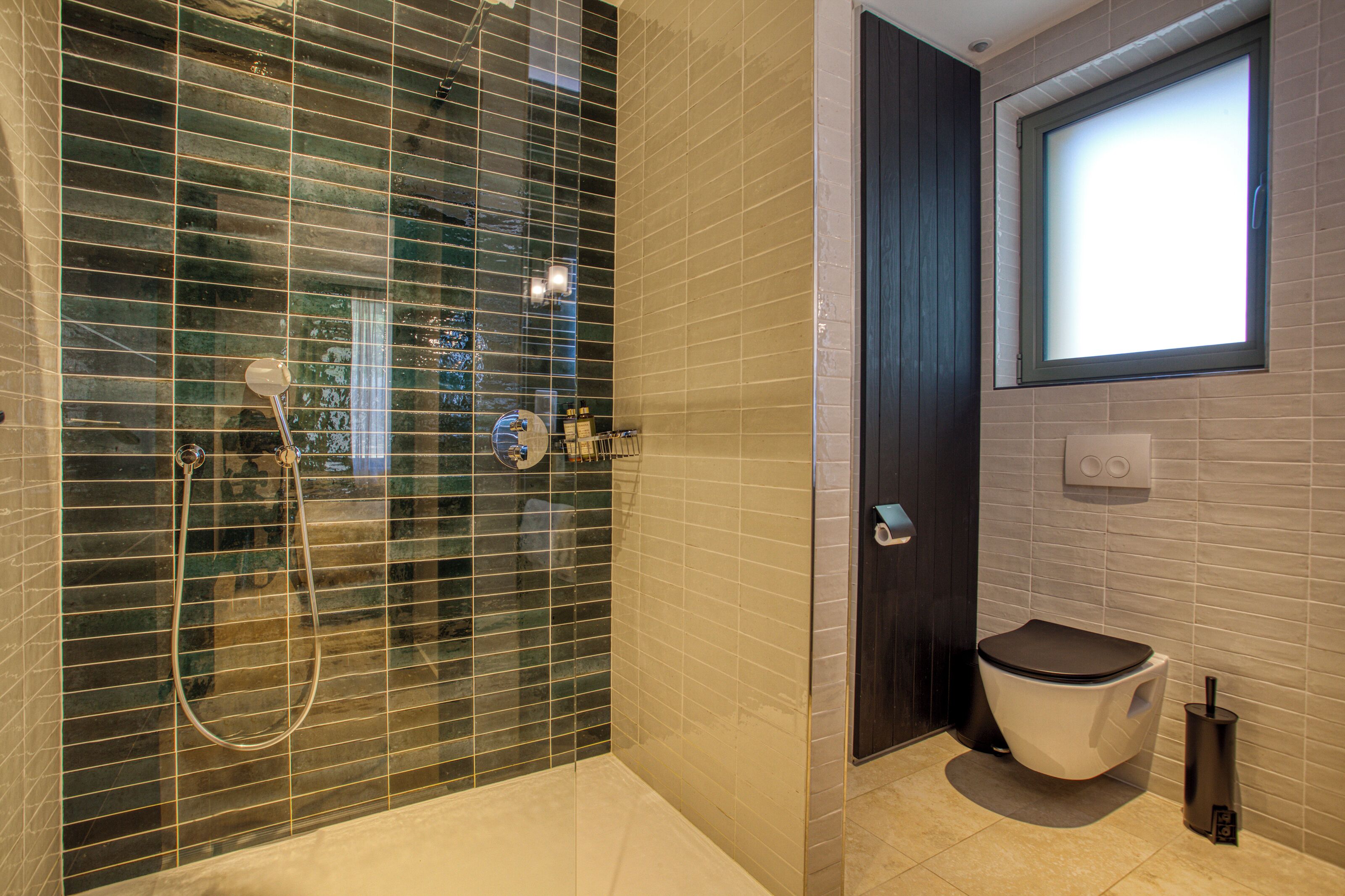 Et moderne badeværelse med et glasafskærmet brusebad, toilet og et vindue.