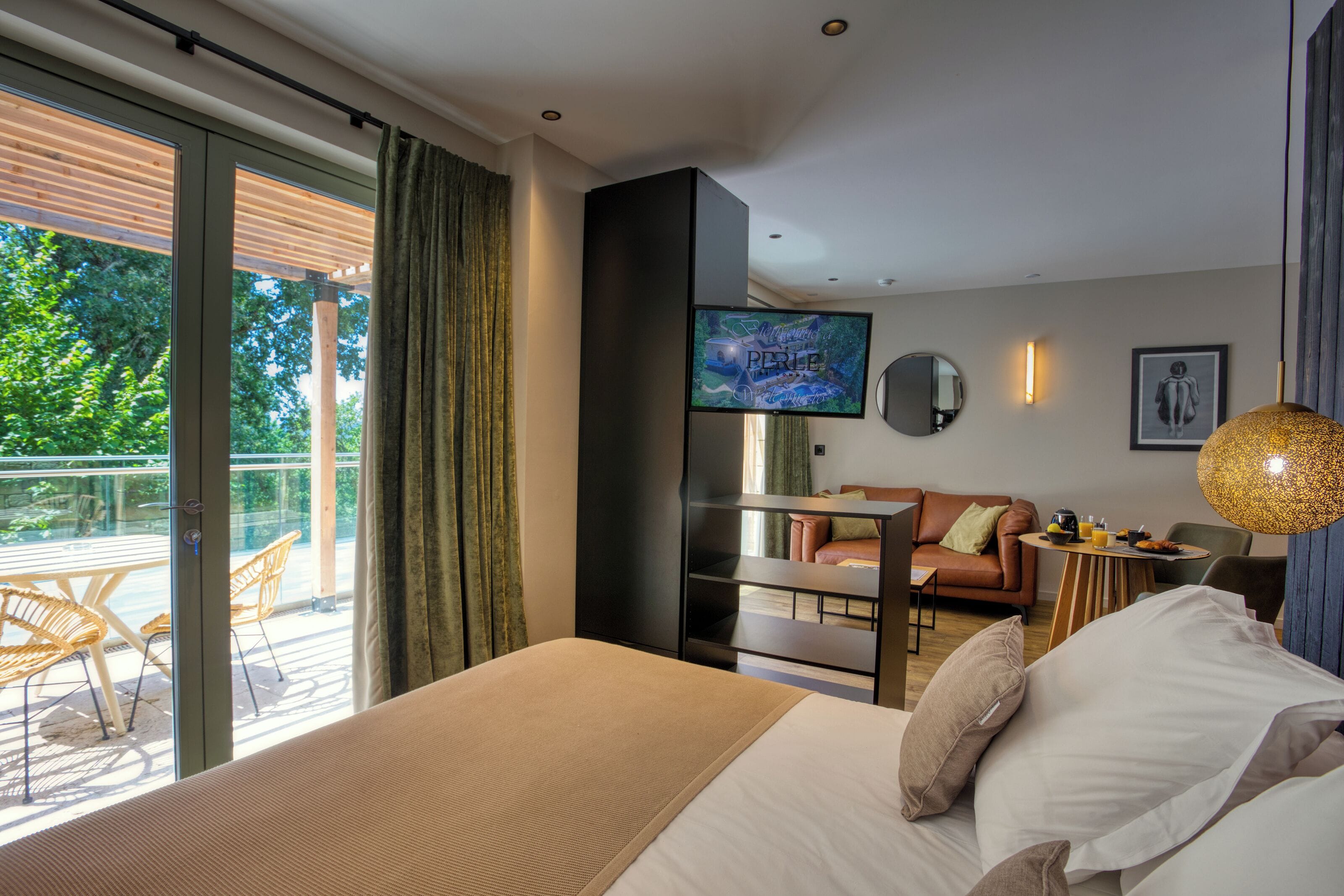 A modern hotel room with a bed, a TV, a sofa, a small table, and a balcony with chairs.