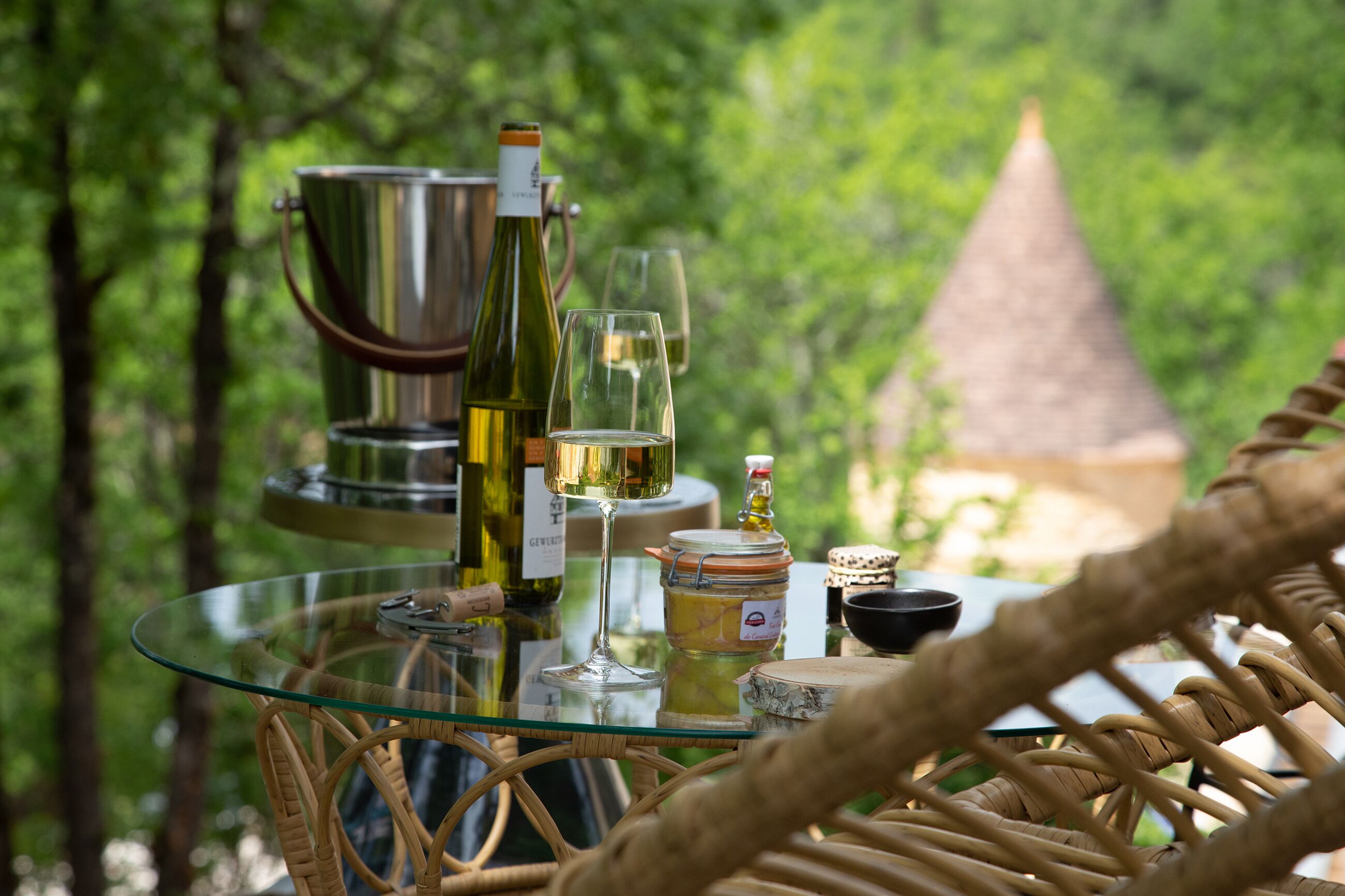 A glass table with bottles, glasses, and a jar in an outdoor setting with greenery in the background.