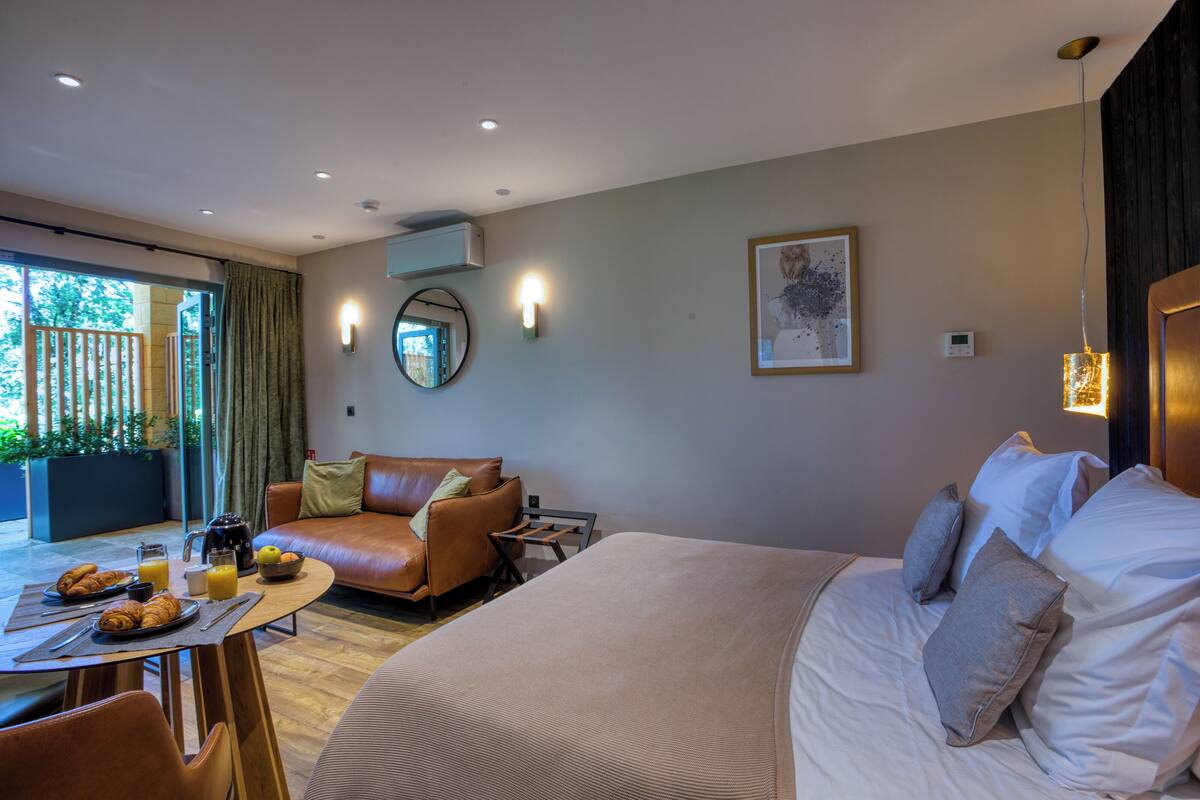 A hotel room with a large bed, a sofa, a dining table with breakfast, and a view of greenery outside.