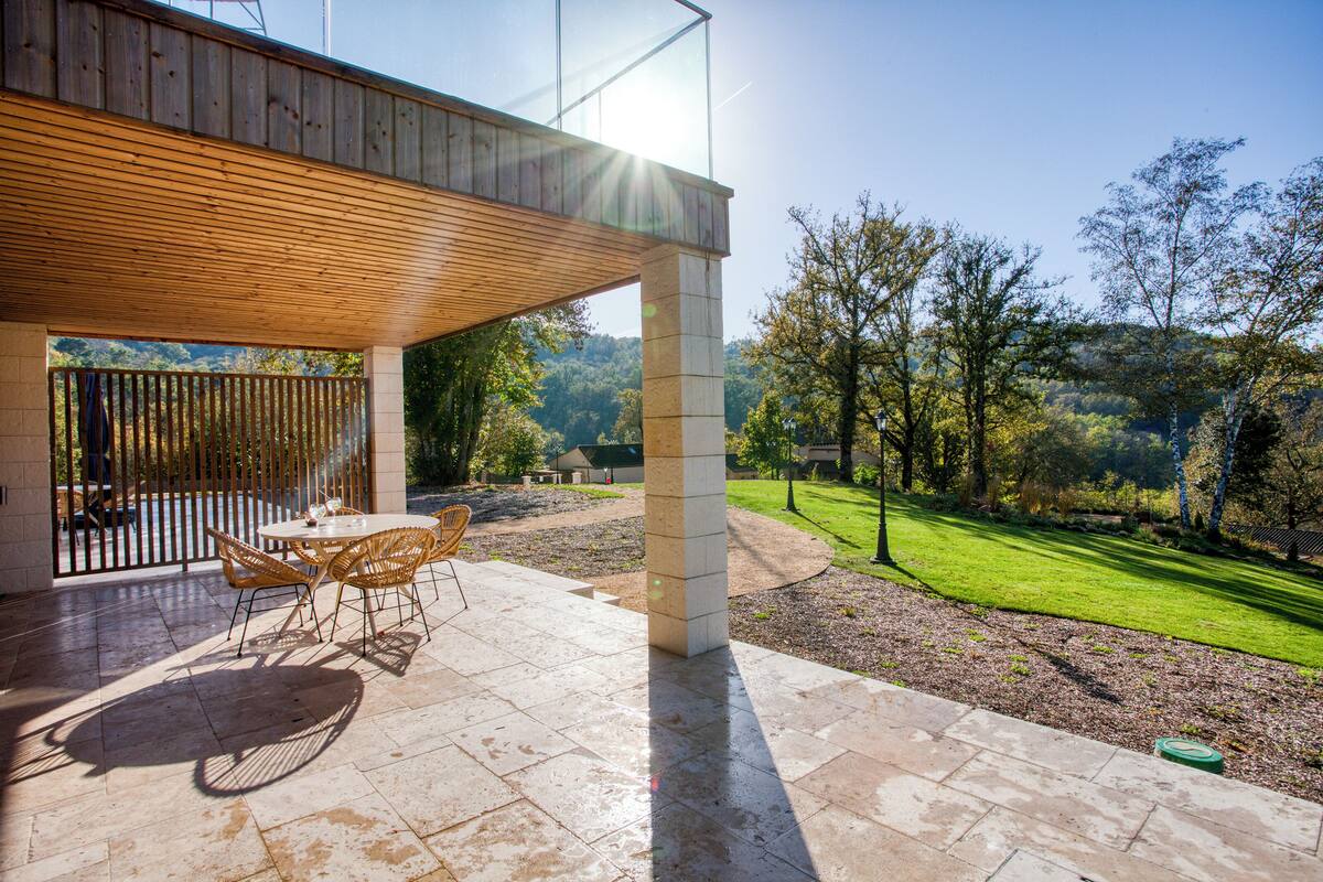 An outdoor patio with a wooden ceiling, a round table with chairs, and a view of a green landscape with trees and a clear sky.