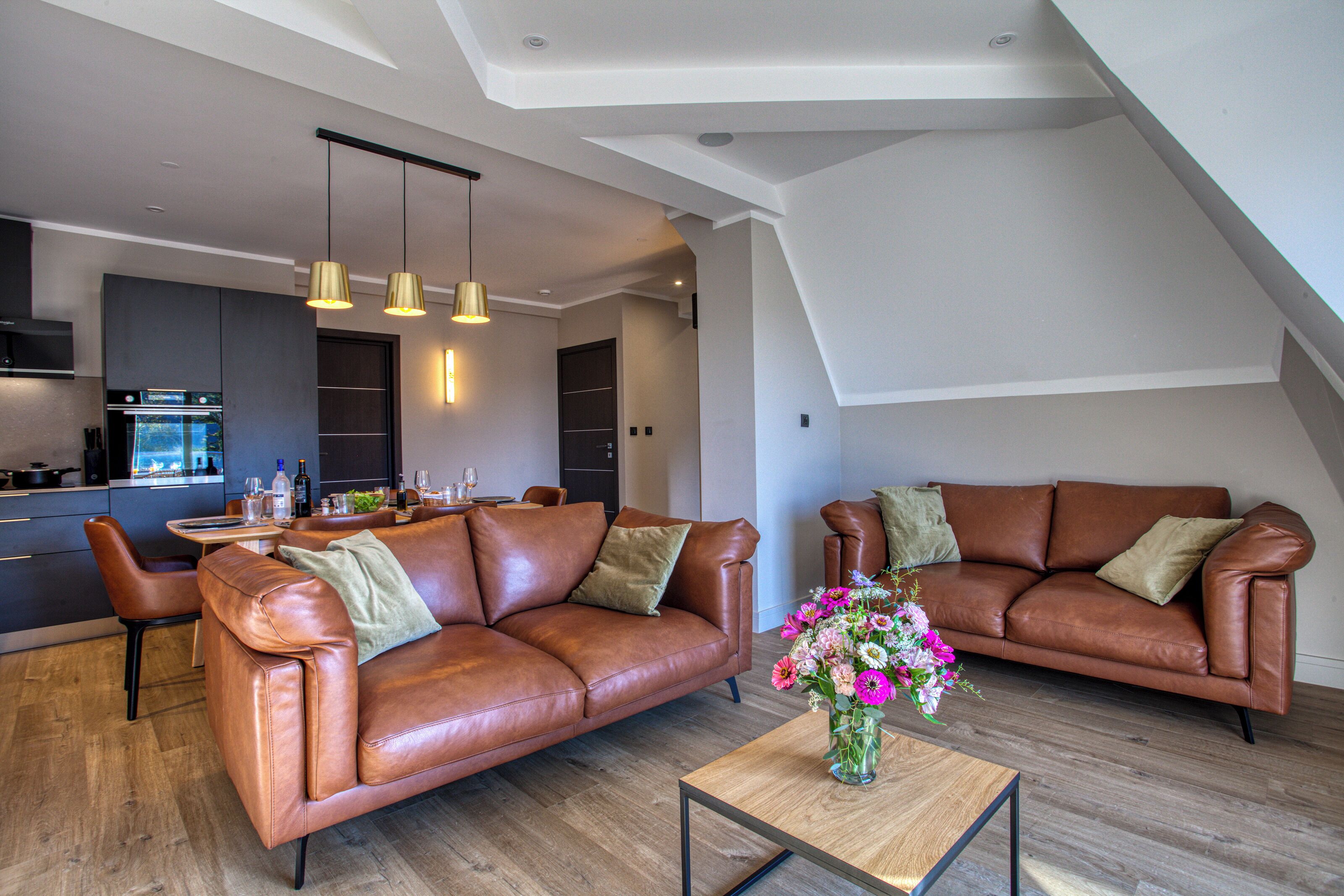 A modern living room with two leather sofas, a wooden coffee table, and a dining area with a kitchen in the background.