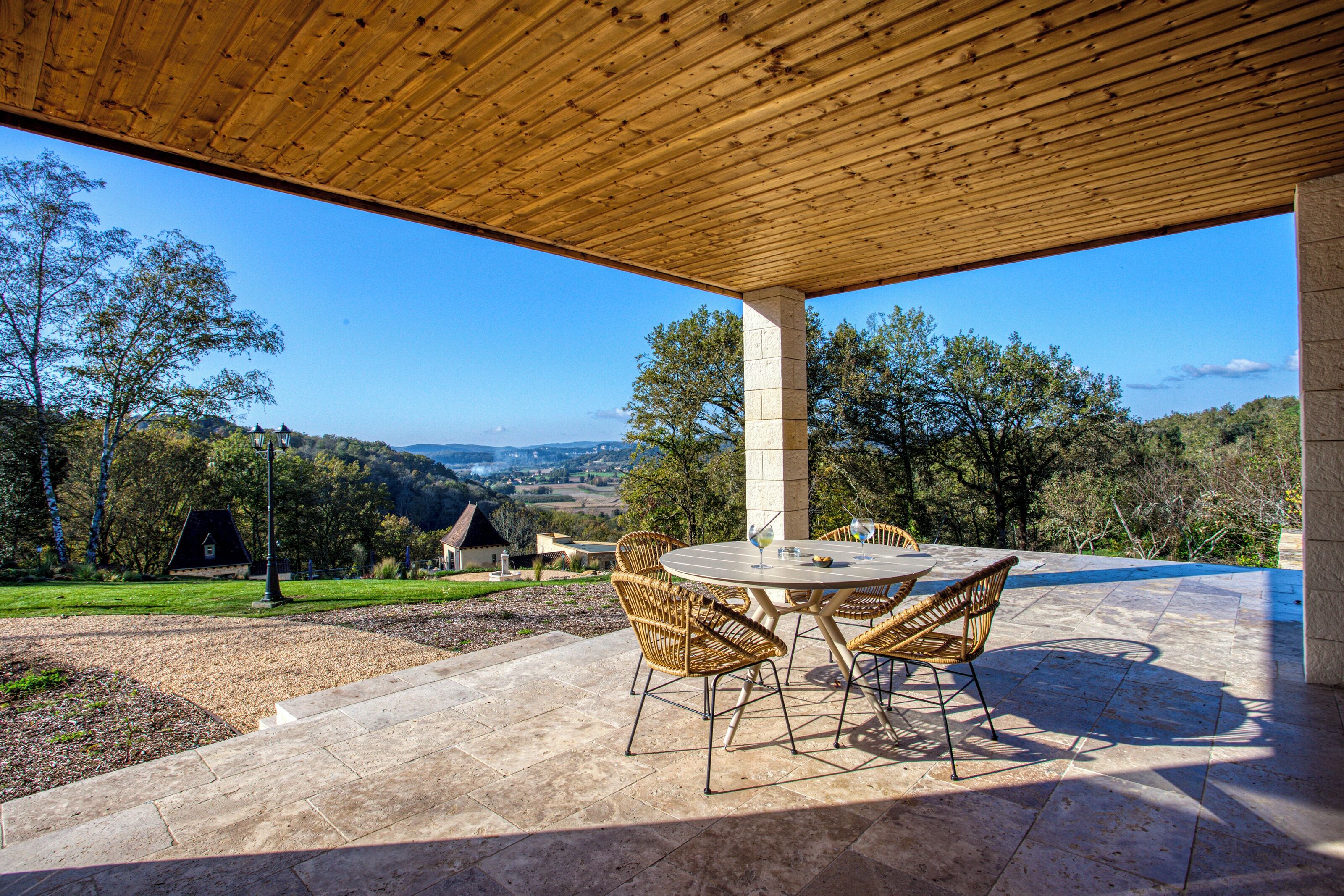 Un patio al aire libre con techo de madera, una mesa redonda con sillas y vista al campo.