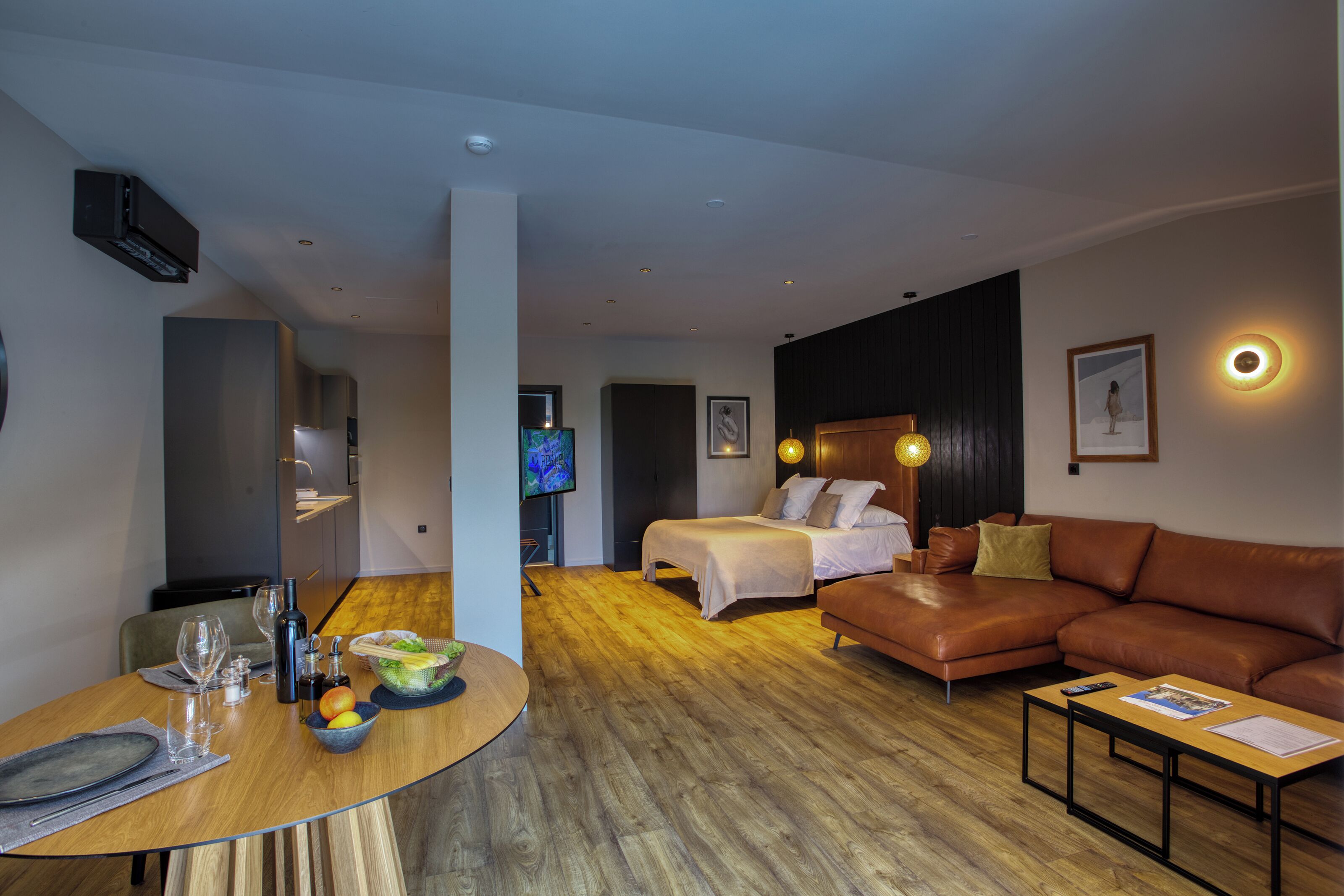 Una habitación de hotel moderna con piso de madera, una cama, un sofá de cuero marrón, una mesa de comedor con vasos y un frutero, y un televisor al fondo.