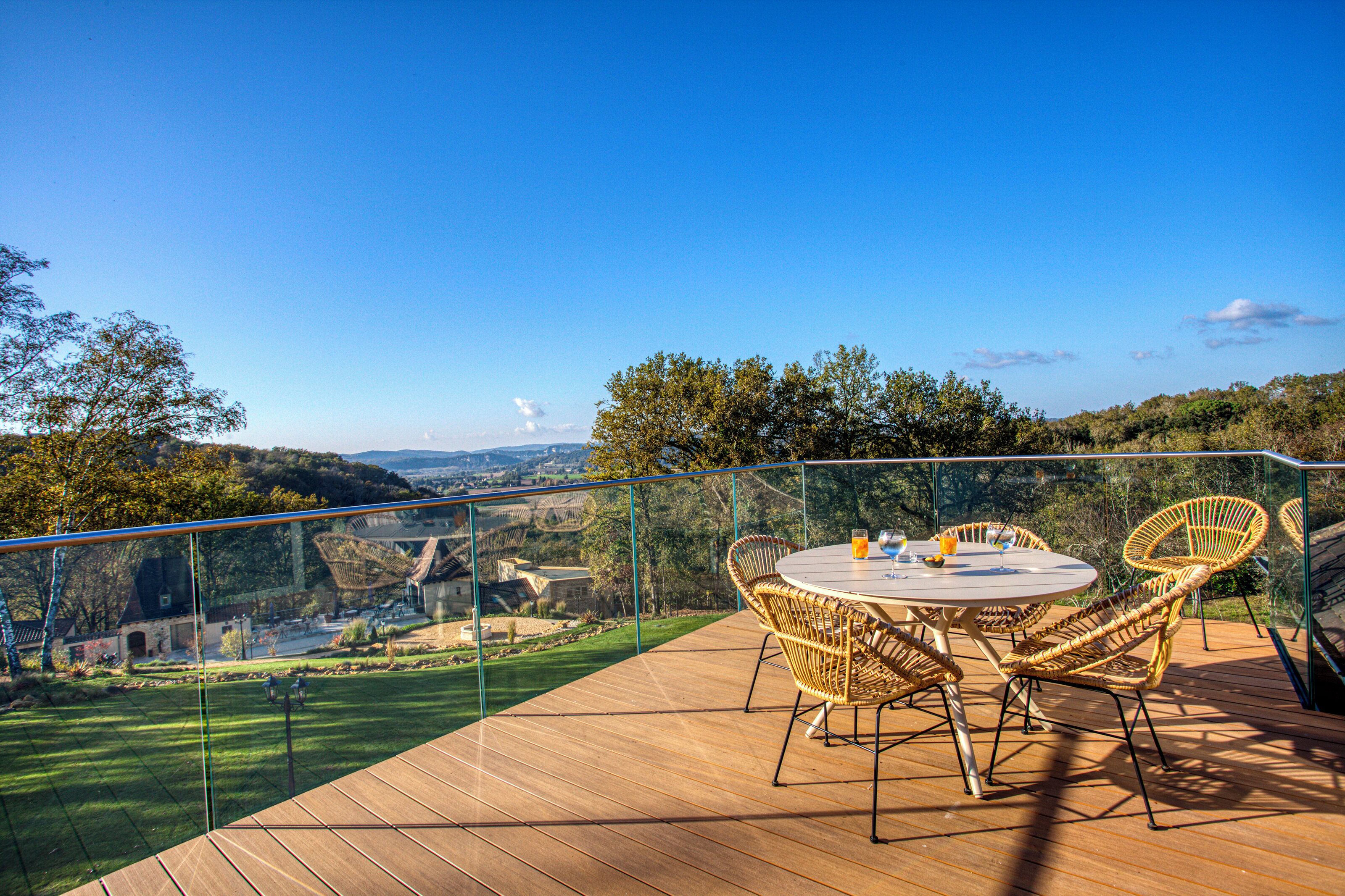 A wooden deck with a glass railing, wicker chairs, and a round table with a glass of orange juice.