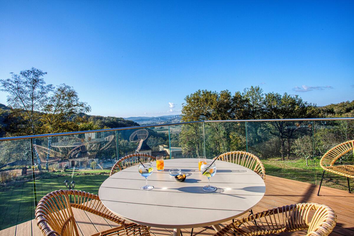 A round table with wicker chairs on a wooden deck, overlooking a scenic landscape with hills and trees.