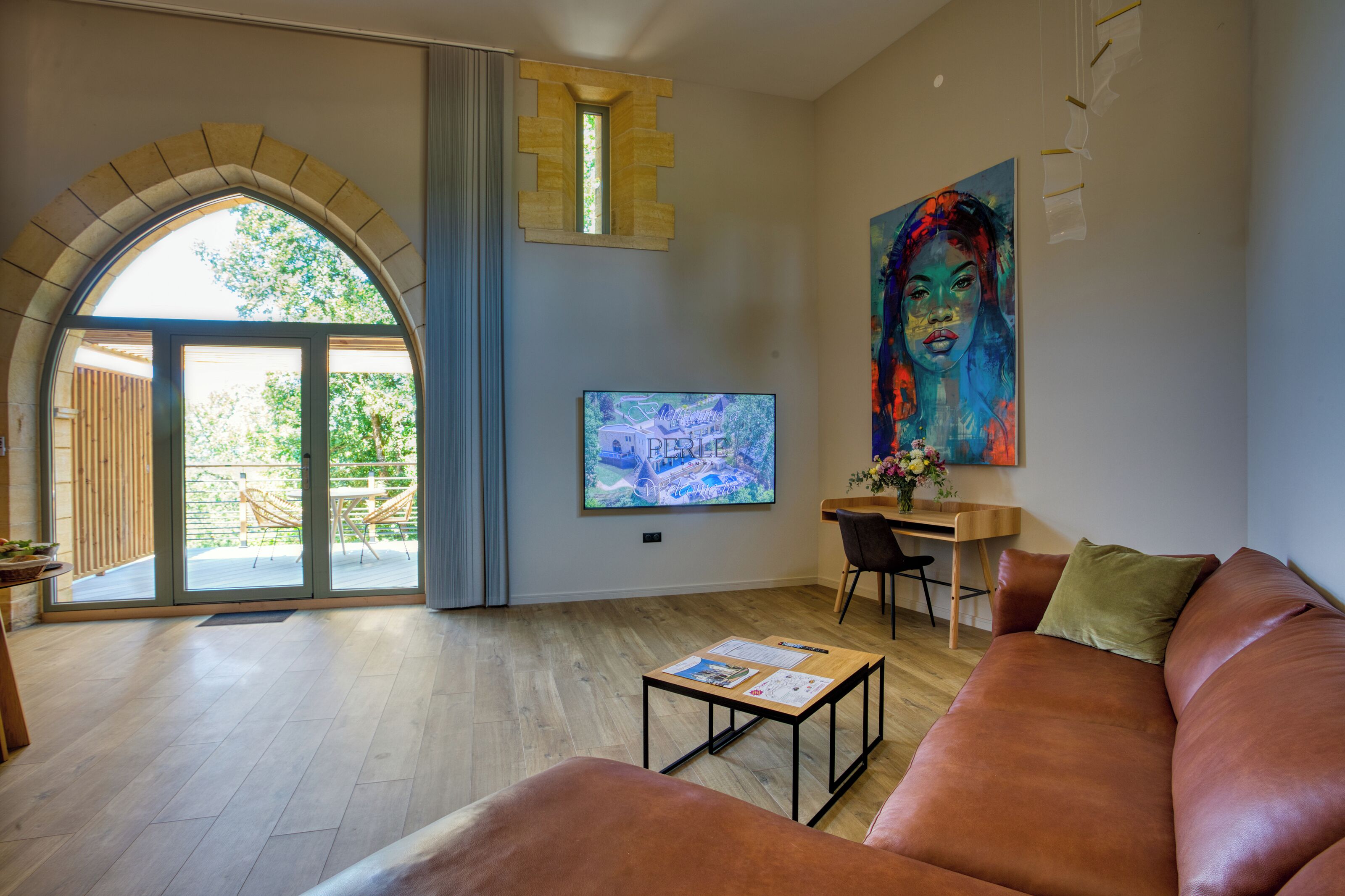 A modern living room with a large arched window, a flat-screen TV, a colorful painting, and a leather sofa.