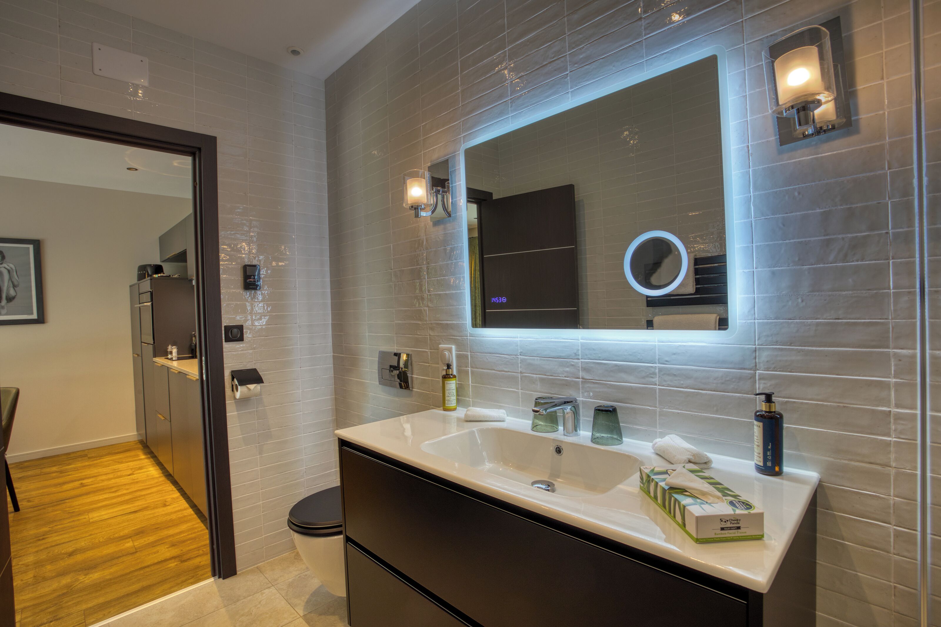 A modern bathroom with a large mirror, a sink, and a toilet. There are toiletries on the sink, and the walls are tiled.
