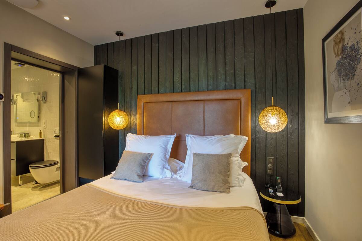 A modern hotel room with a large bed, two bedside lamps, a small table with a phone, and a view of the bathroom.
