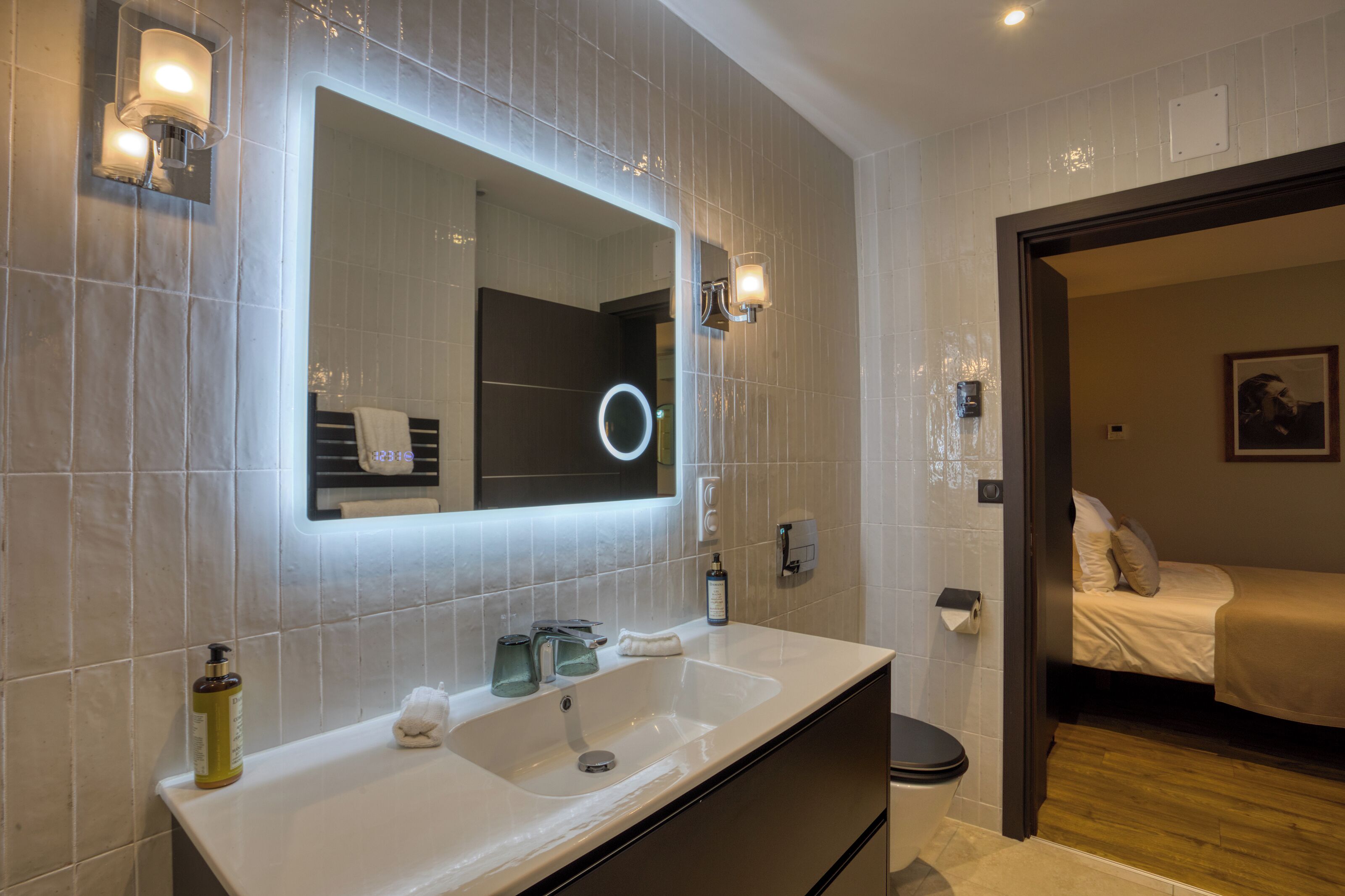 A modern bathroom with a large mirror, a sink, and a toilet. There is a small bedside table with a lamp and a bottle of lotion.