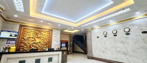 Zona de recepción de un hotel con un mostrador de mármol, un panel decorativo en la pared, una escalera y un reloj.