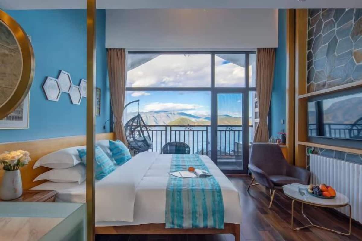 A hotel room with a bed, a chair, a small table with fruit, and a view of the mountains through large windows.