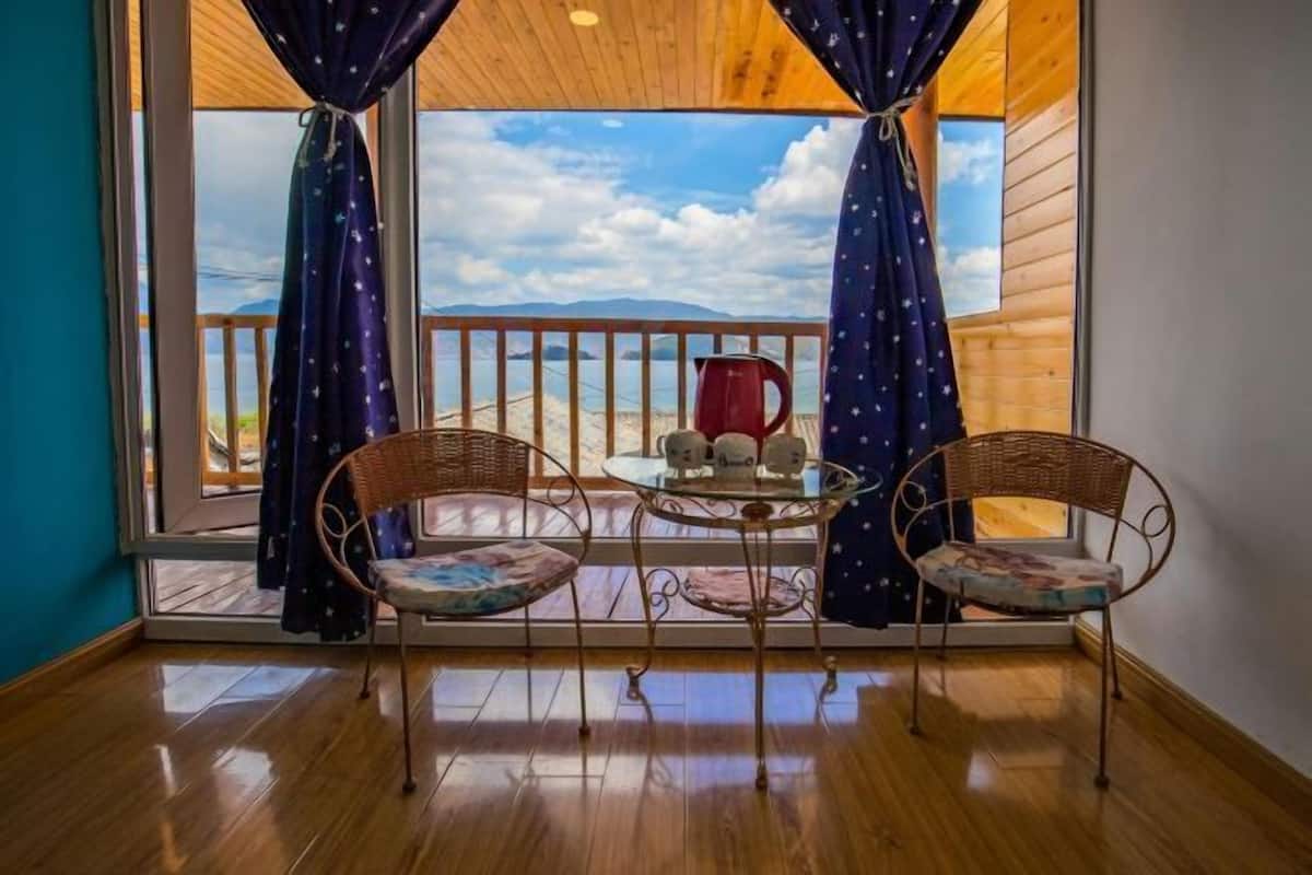 A balcony with wicker chairs and a small round table, a red kettle, and a scenic view of a lake and mountains.