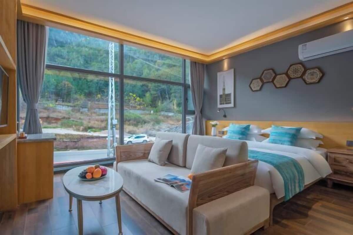 A modern hotel room with a large window, a sofa, a small table with fruit, and a bed with a blue and white bedspread.