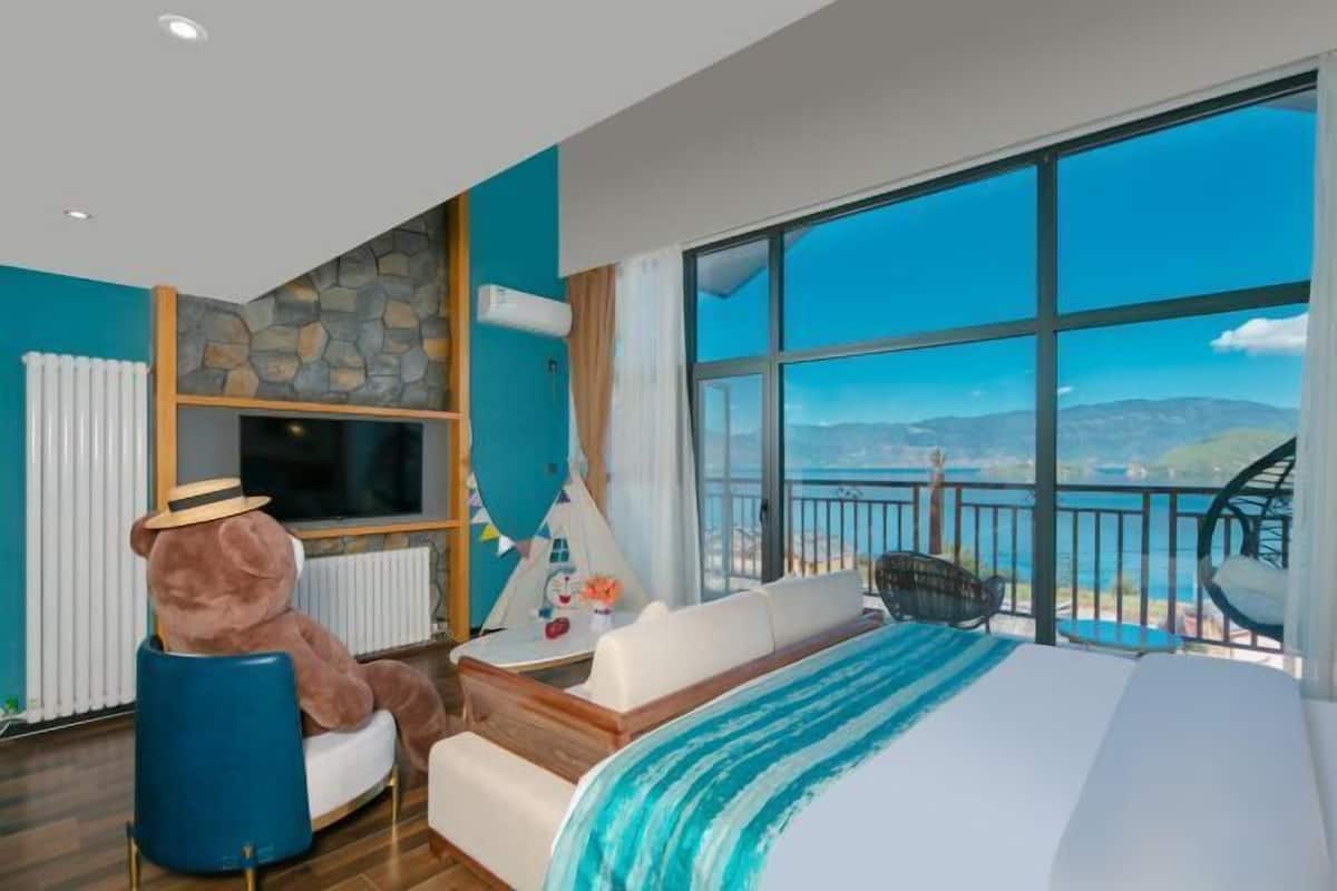 A hotel room with a large bed, a view of the sea, a stone wall, and a teddy bear.