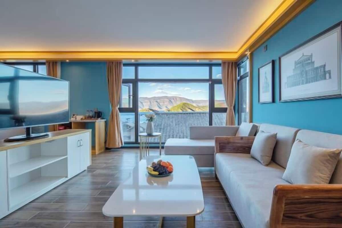 A modern living room with a flat-screen TV, a white coffee table with fruit, a sofa, and a large window with a mountain view.