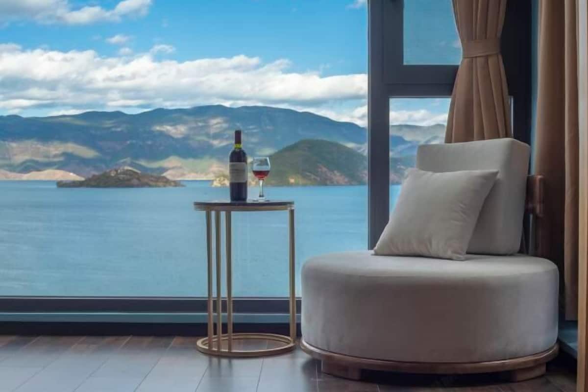 A room with a large window offering a view of a lake and mountains. There is a round chair and a small table with a bottle of wine and a glass.