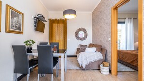 Une pièce de taille modeste comprenant une table à manger, des chaises, un lit, un petit tapis, un miroir fixé au mur et une porte donnant sur une autre pièce.