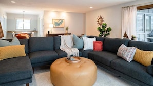 Ein modernes Wohnzimmer mit einem dunkelgrauen Ecksofa, einem runden Hocker und einem Essbereich im Hintergrund.