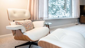 Ein modernes Wohnzimmer mit einem weißen Ledersofa, einem passenden Ottomanen und einem kleinen runden Tisch. Ein großes Fenster mit Vorhängen bietet Blick auf schneebedeckte Bäume.