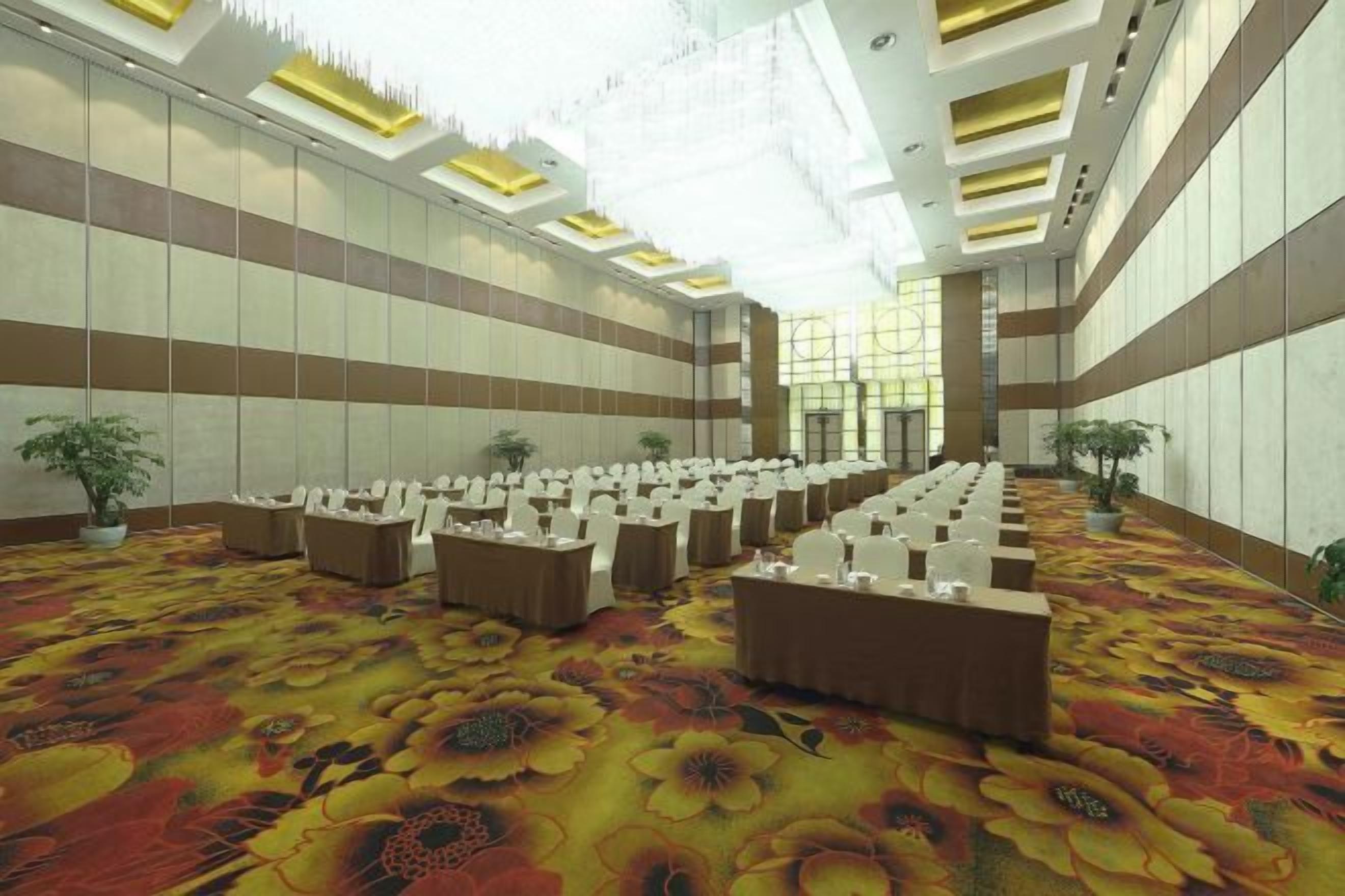 Un amplio salón de conferencias con filas de mesas y sillas, alfombra estampada y un techo alto con tragaluces.