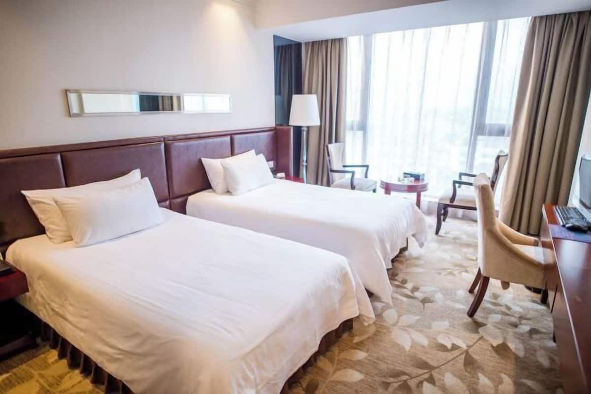 A hotel room with two beds, a desk, a chair, a lamp, and a large window with curtains.