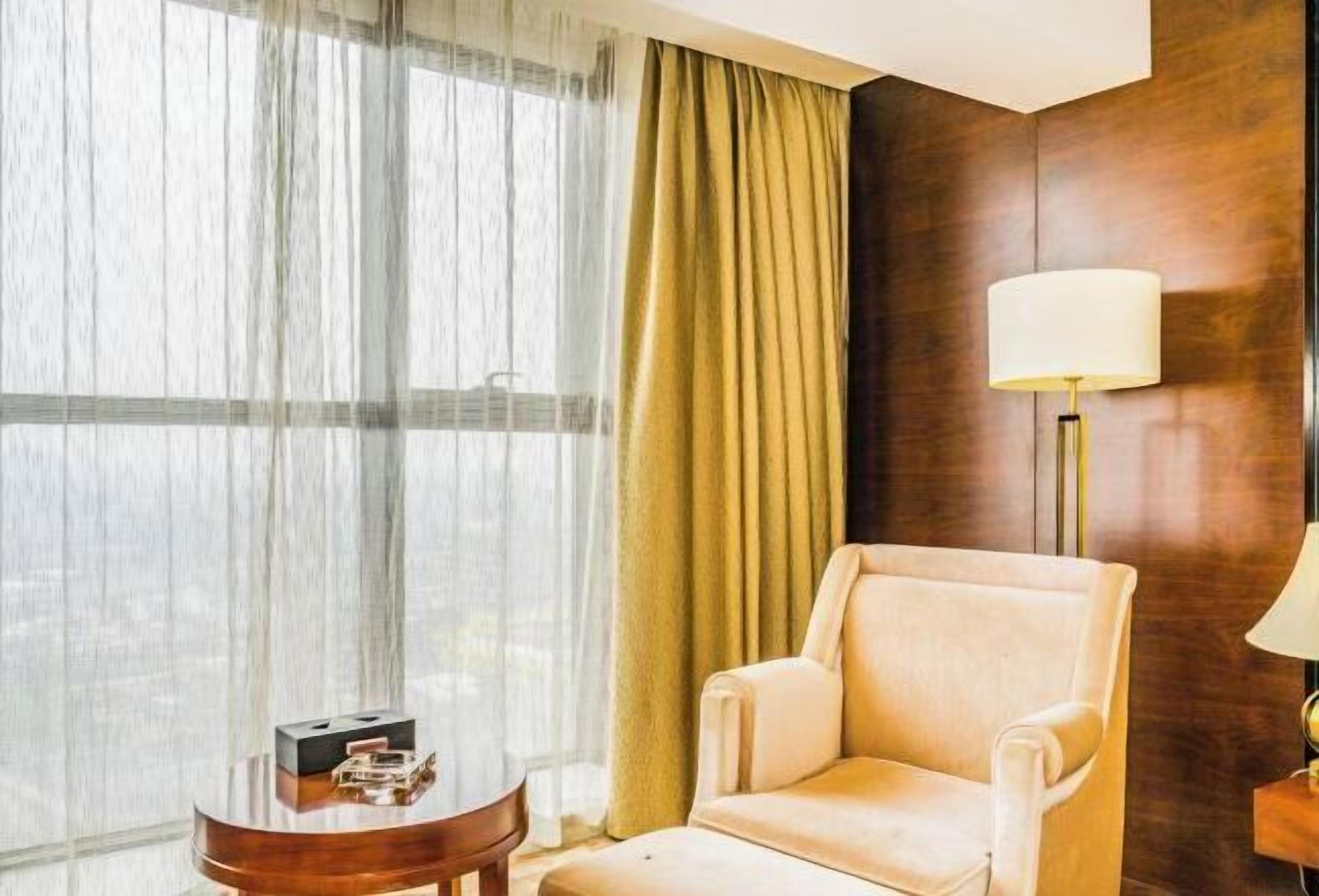 Habitación de hotel con un ventanal grande, un sillón color crema, una mesita redonda pequeña y una lámpara de pie.