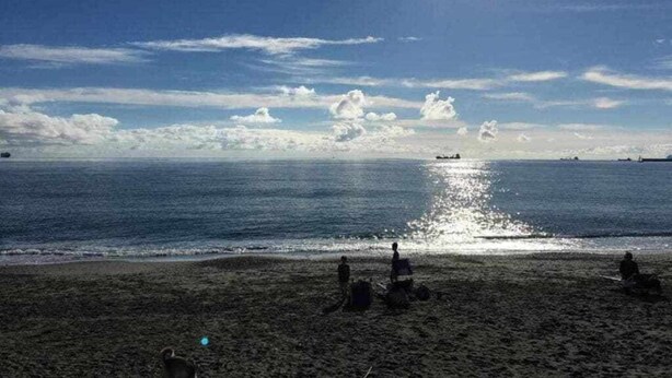 Una spiaggia con persone sedute sulla sabbia, cielo sereno e una nave all'orizzonte.