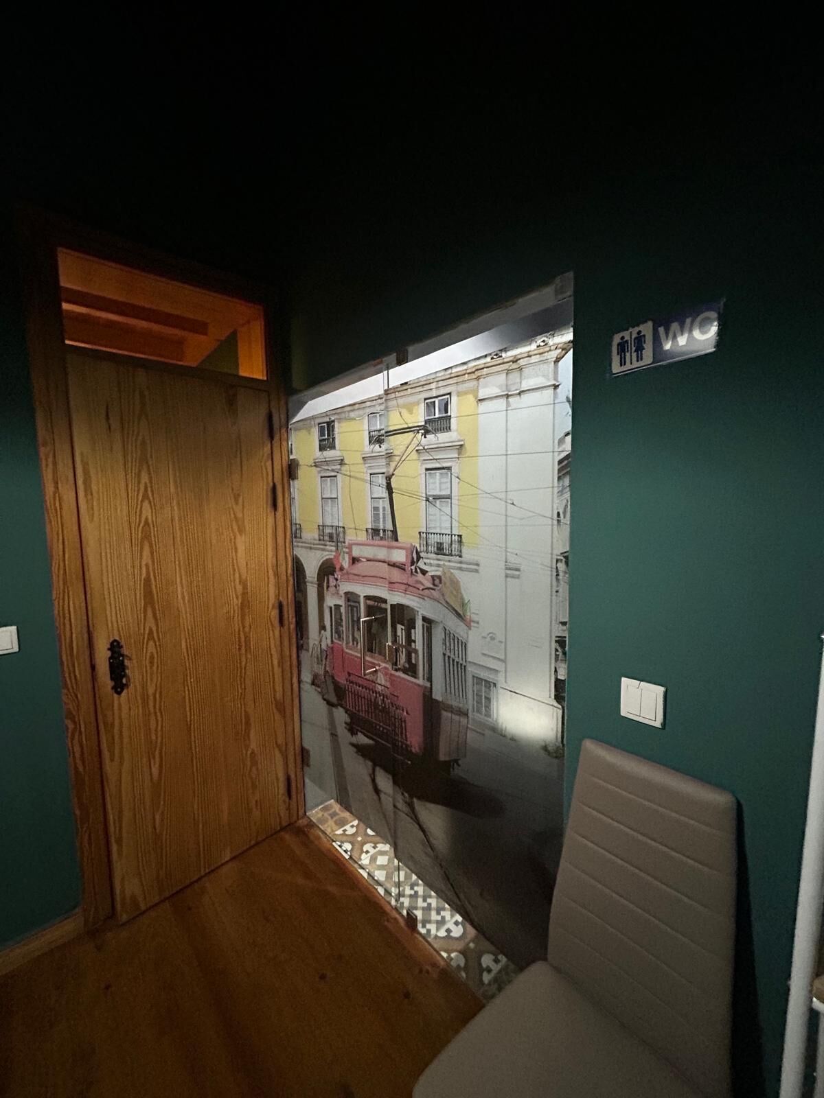 Ein Zimmer mit Holztür, Wandbild einer Straßenszene, WC-Schild und einem Stuhl.