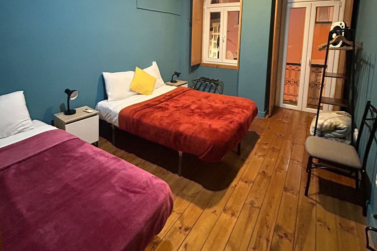 A room with two beds, a wooden floor, a window, and an air conditioning unit.