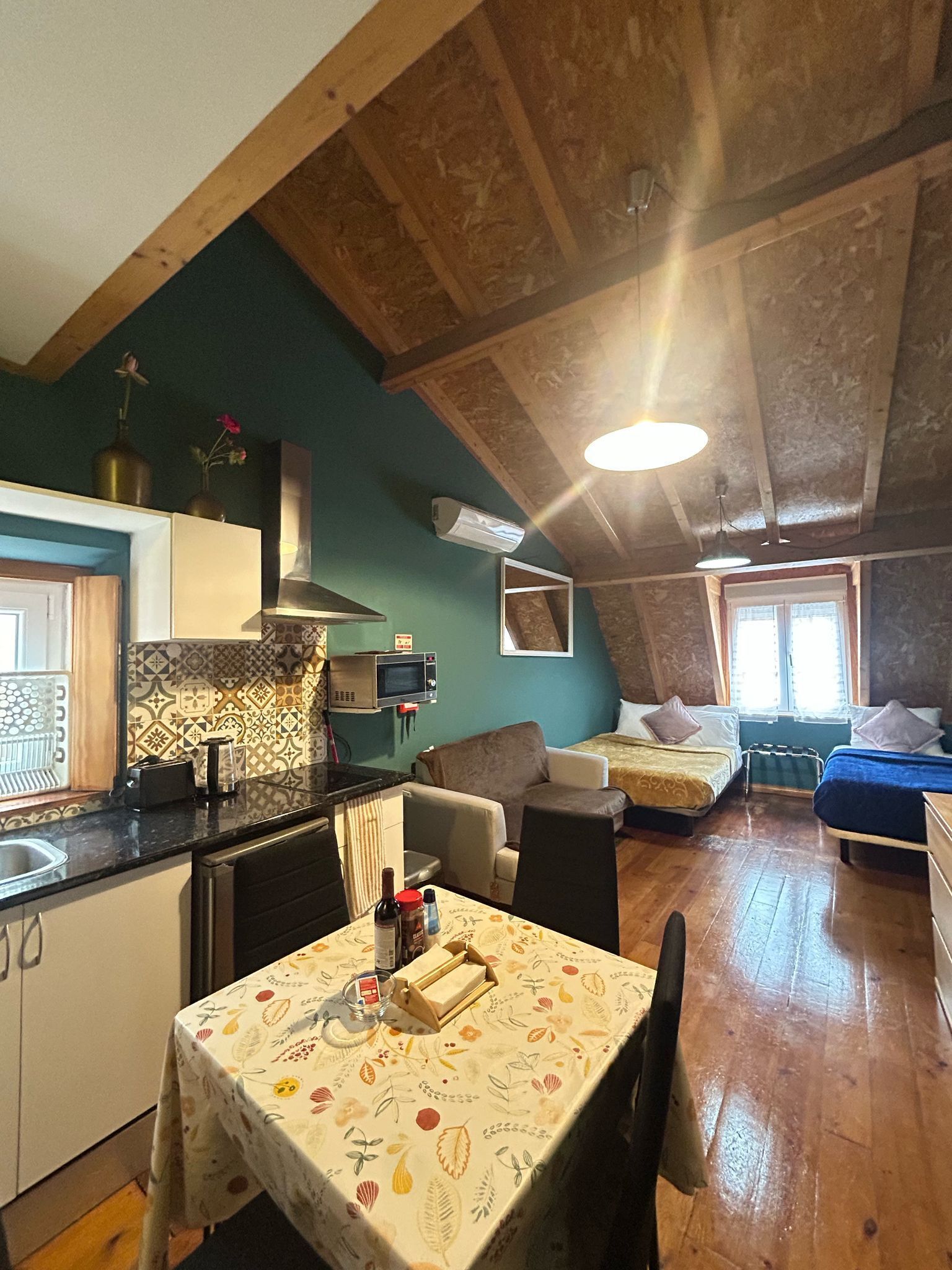 A cozy kitchen with a dining table, a microwave, and a sink. There are two beds with pillows and a window with curtains.