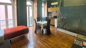 A spacious room with a wooden floor, a dining table with chairs, a red bench, a glass shower enclosure, and a kitchen area with a stove and cabinets.
