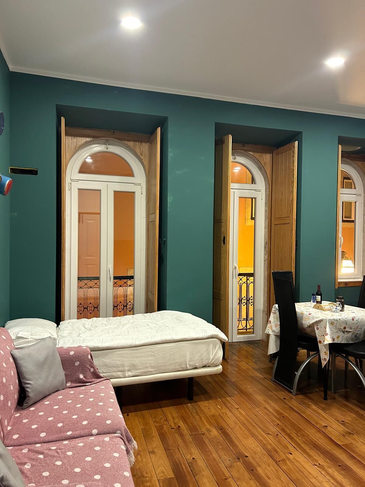 A room with teal walls, a wooden floor, a bed, a sofa, a dining table with chairs, and two doors leading to an outdoor area.