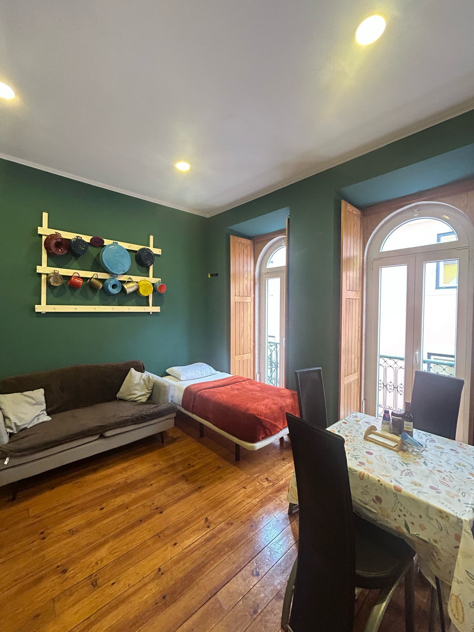 A room with a wooden floor, a bed with a red blanket, a sofa with pillows, a table with a floral tablecloth, and a wall-mounted shelf with colorful items.