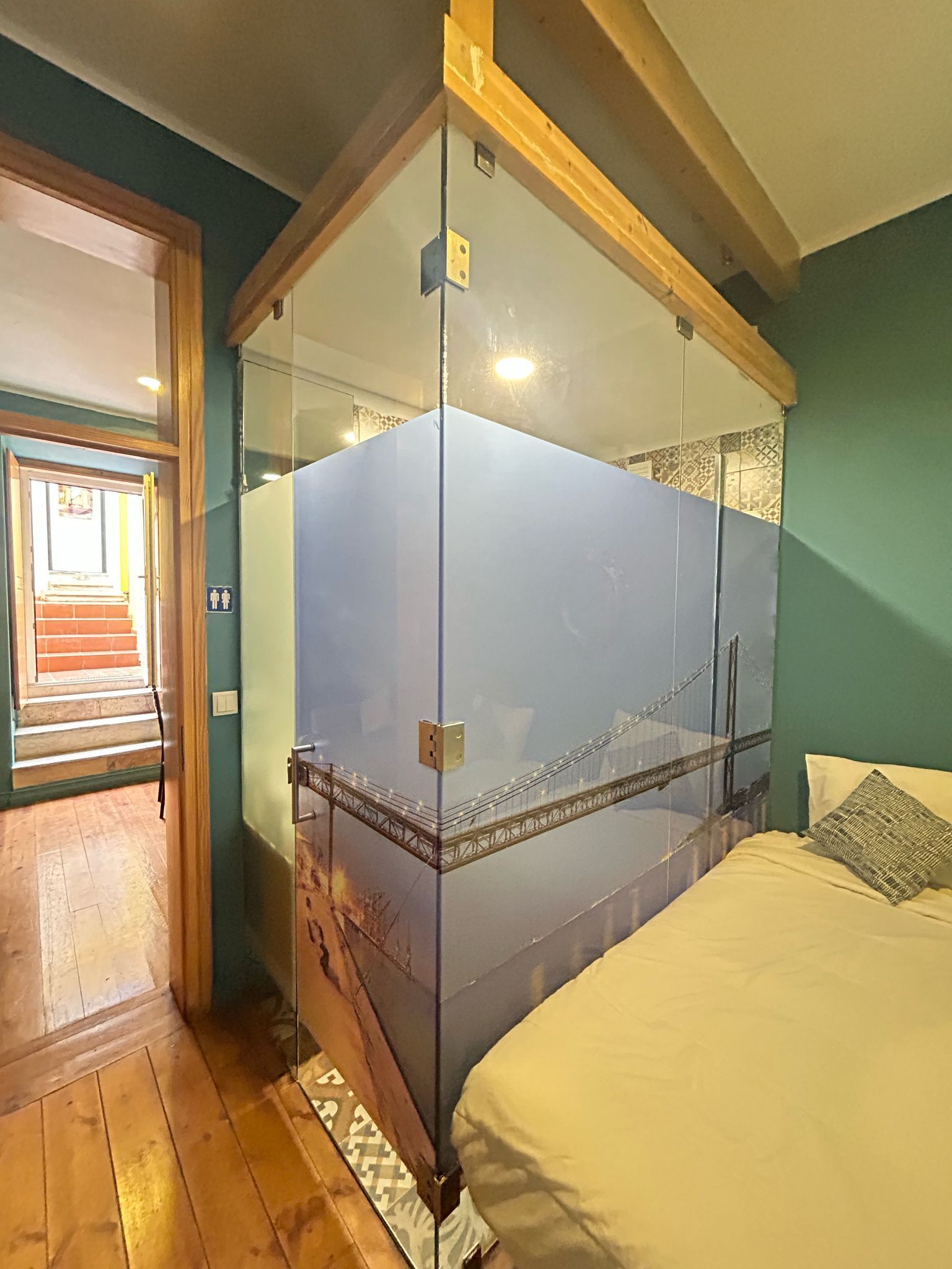 A bedroom with a glass partition bed, a wooden floor, a bed with white bedding, and a view of a bridge through the glass.