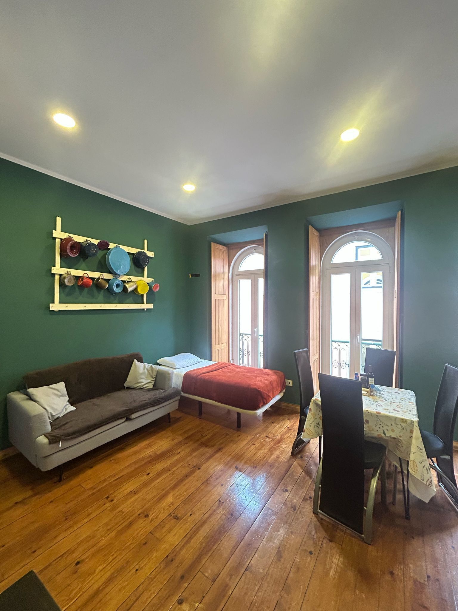 A room with a wooden floor, a sofa, a bed, a dining table with chairs, and a wall-mounted shelf with colorful items.