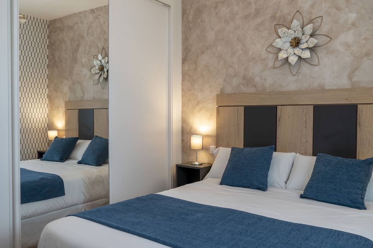 A hotel room with two beds, a wooden headboard, a wall-mounted flower decoration, and a nightstand with a lamp.