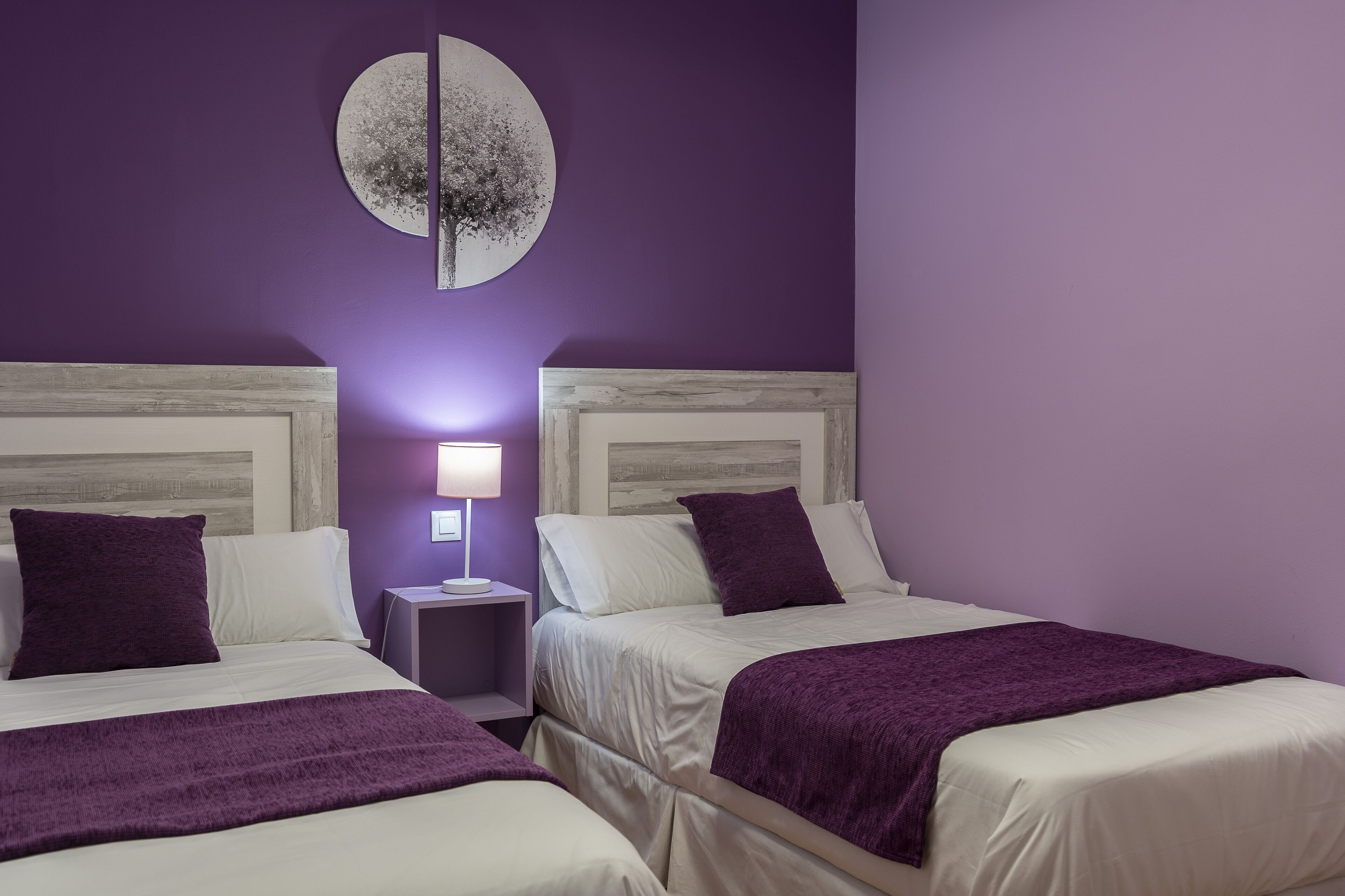 A room with two beds, each with white bedding and purple accents, a nightstand with a lamp, and a wall-mounted circular artwork.